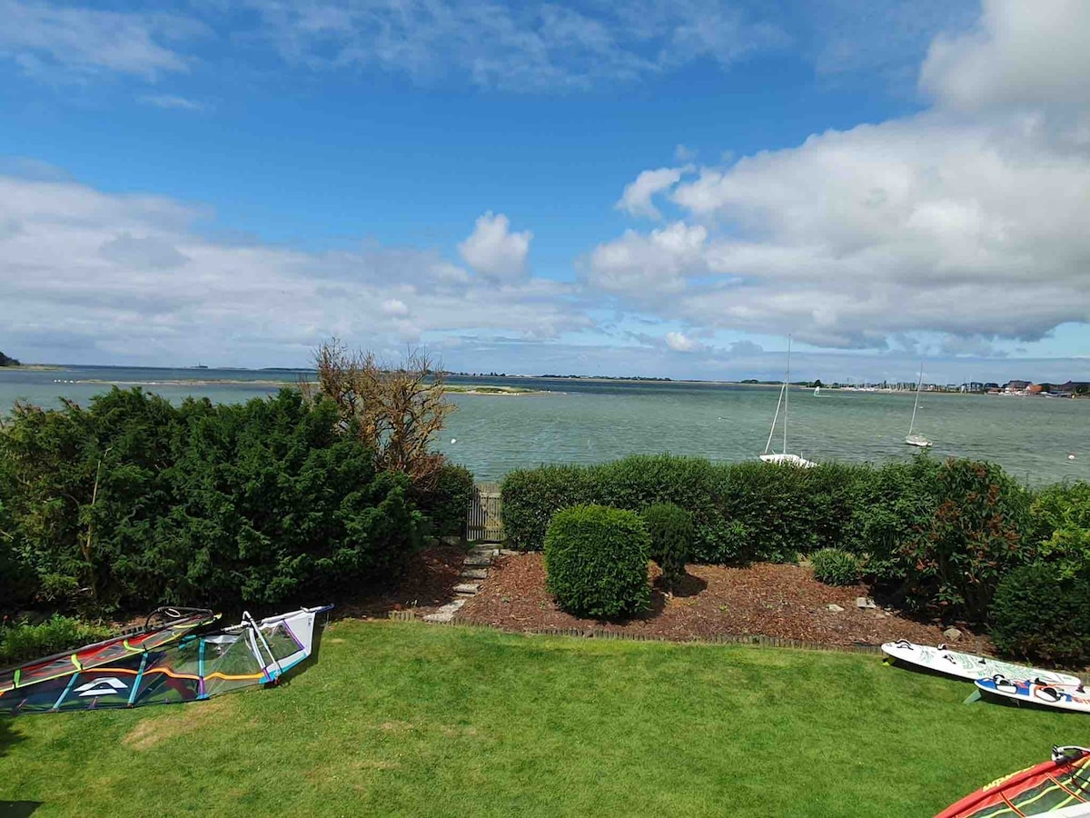 A view of the serene waterfront is presented, featuring a grassy area with sailing equipment and shrubs. The water reflects the cloudy sky, with boats visible in the distance. The landscape is framed by greenery, providing a peaceful coastal atmosphere.