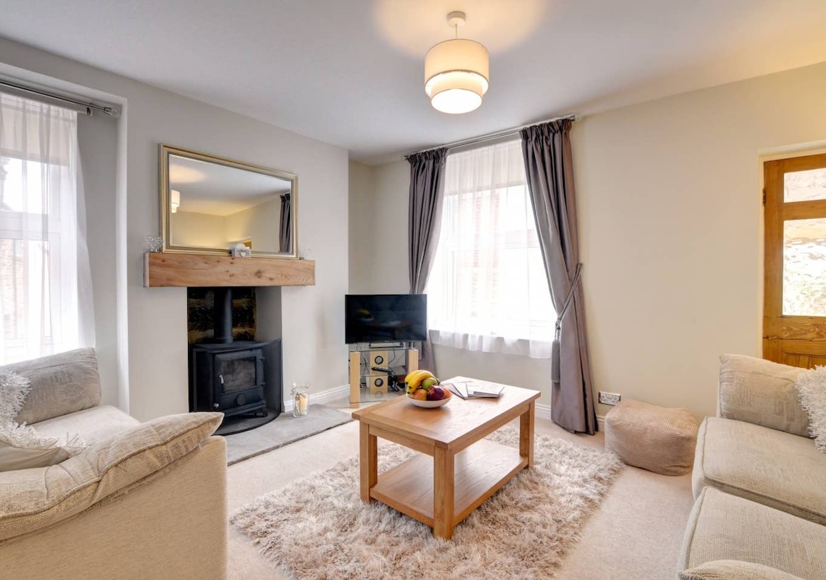 A comfortable living area is presented with two soft sofas and a central wooden coffee table. A log burning stove is situated beneath a decorative mirror, complemented by a TV. Large windows allow natural light to fill the space, which features neutral tones and gentle textures.
