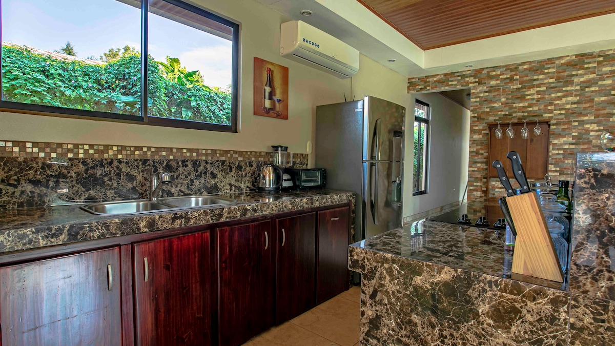 The kitchen features dark wood cabinetry and a striking granite countertop. Modern appliances, including a refrigerator and coffee maker, are visible. Large windows allow natural light to fill the space while offering a view of the greenery outside.
