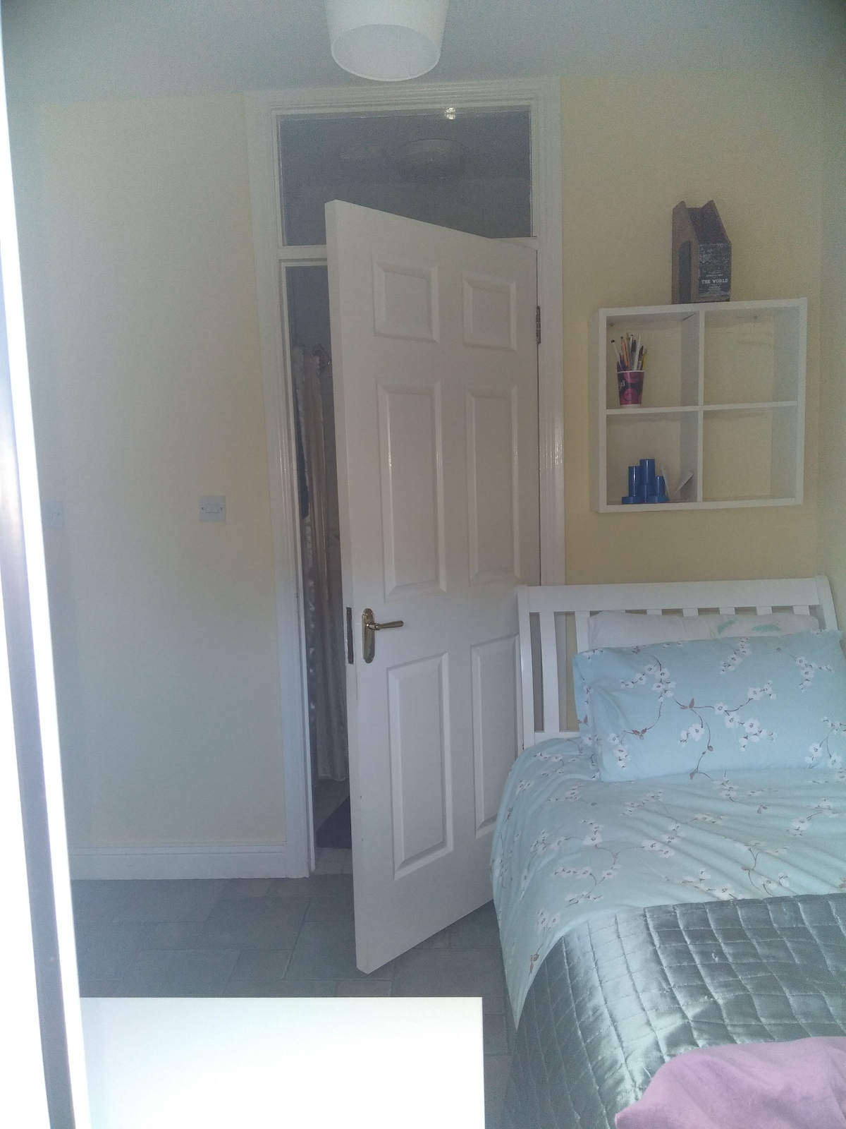 A cozy bedroom features soft-toned walls and a neatly made bed with a light-colored comforter. A door leads to an ensuite wetroom, and a white shelf displays various toiletries. Light from a nearby window enhances the inviting atmosphere of the space.
