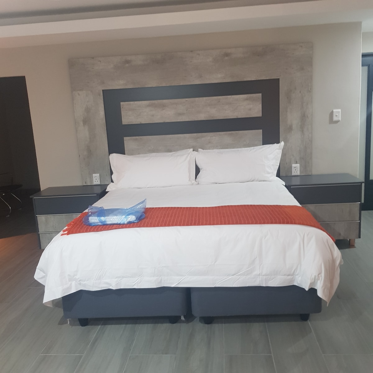 A spacious bed is positioned against a modern headboard featuring a textured design. Each side of the bed is flanked by minimalistic nightstands. Crisp white bedding is complemented by an orange throw neatly placed at the foot of the bed. Natural light is welcomed through a nearby window.