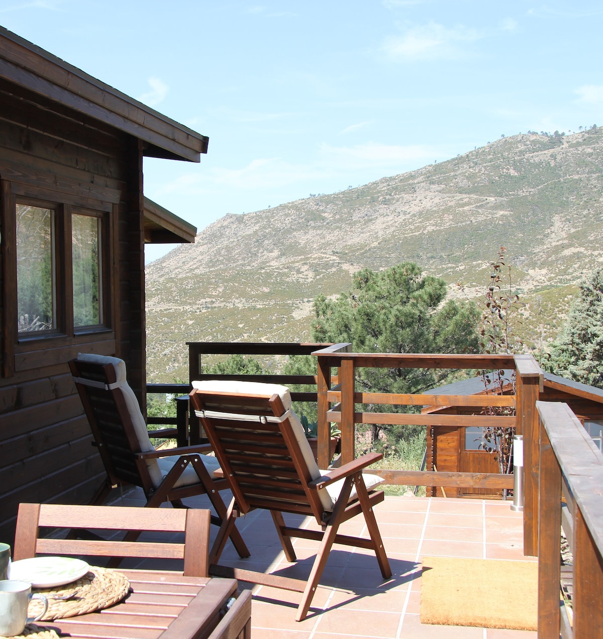 A wooden deck features two folding chairs overlooking a scenic mountain view. The surrounding landscape includes a mixture of greenery and rocky terrain under a clear blue sky, providing a peaceful outdoor setting.