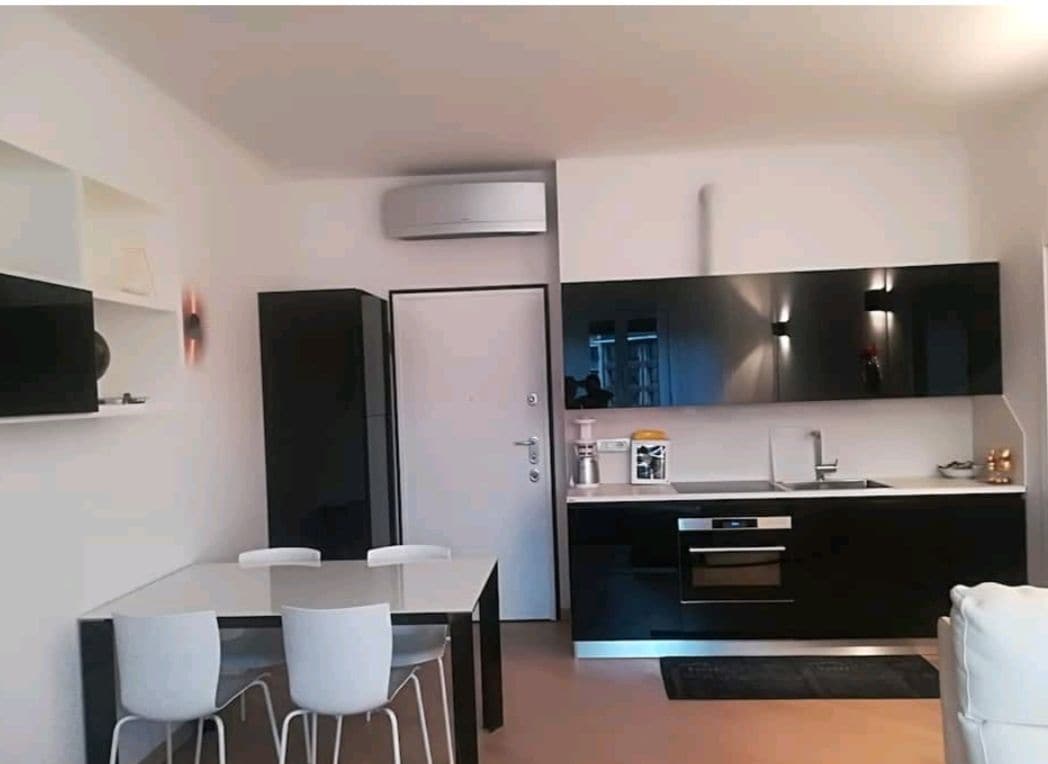 An open space kitchen is showcased, featuring sleek black cabinetry and modern appliances. A dining table with four white chairs is positioned near the window. A comfortable sofa is visible in the corner, contributing to the inviting layout of the area.