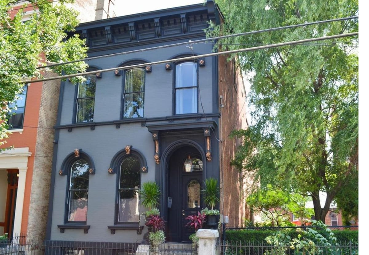 A historic two-story mansion is displayed in a classic style, showcasing detailed architectural features and large arched windows. Lush greenery surrounds the exterior, enhancing the building's charm. A decorative gate leads to the beautifully landscaped front garden.