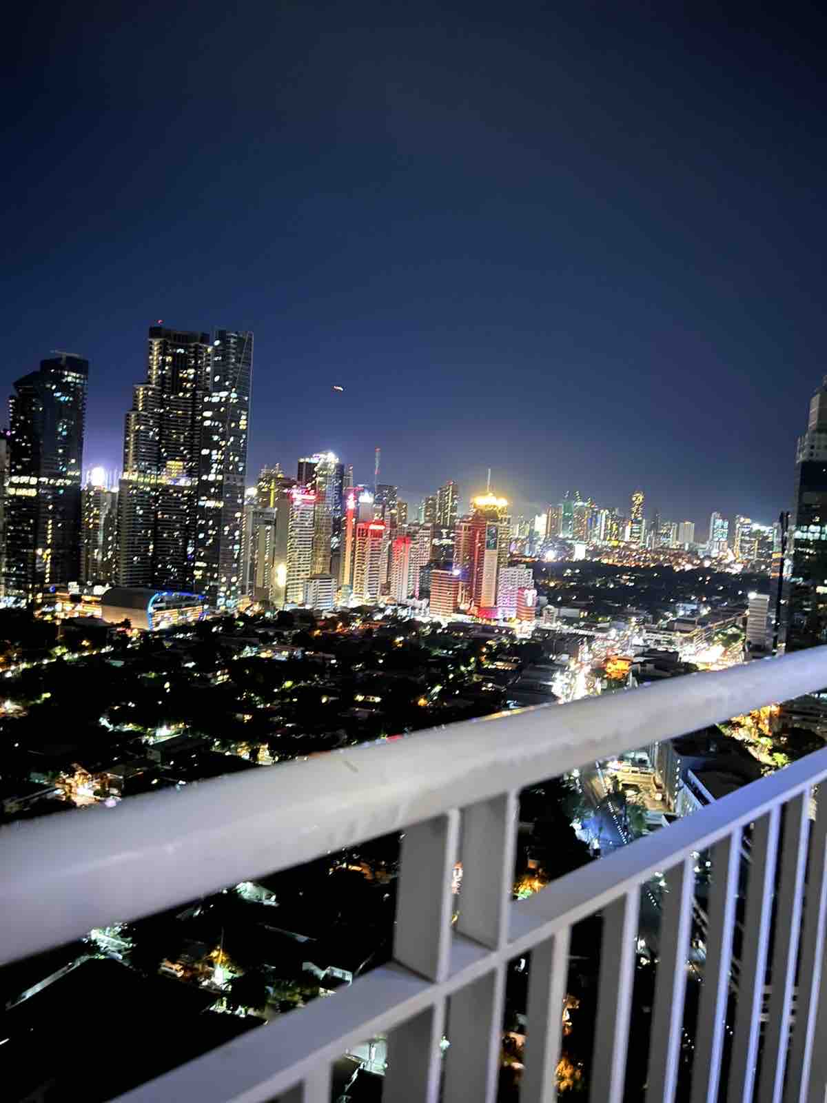 A panoramic view from the balcony showcases a vibrant city skyline illuminated at night. Tall buildings reflect various colors of light, while the streets below are dotted with warm glows, creating a lively urban atmosphere.