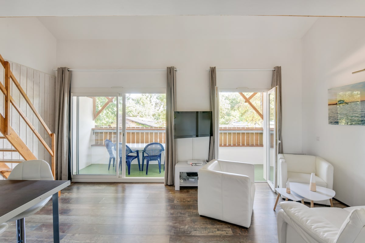 A light-filled living area features a comfortable seating arrangement with white furniture. Large glass doors provide access to a private terrace with outdoor seating. Wooden flooring adds warmth, while neutral walls enhance the airy ambiance.