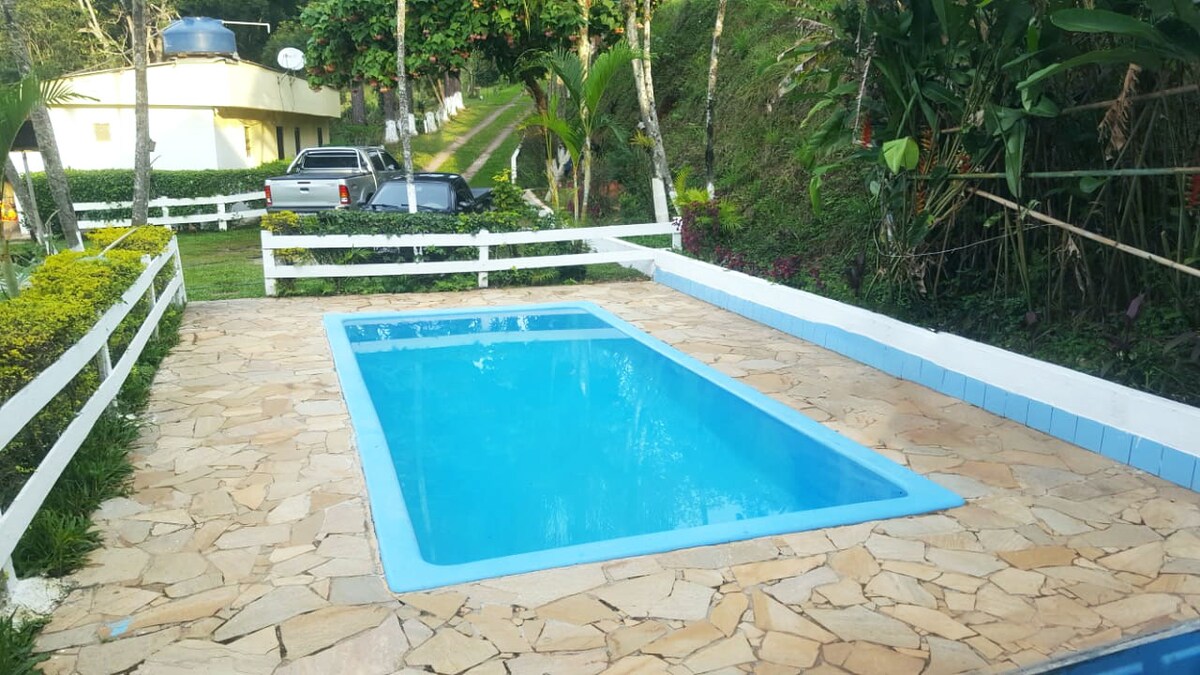 A rectangular swimming pool is surrounded by a stone patio, featuring blue tiles along the water's edge. Lush greenery and colorful plants are visible in the background, with a sloped pathway leading upwards. White fencing borders the area, enhancing the natural setting.