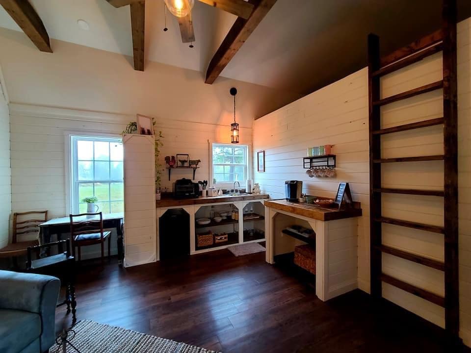 A cozy kitchen nook features wooden beams overhead and bright windows allowing natural light. A desk with a chair is positioned beside a compact countertop, which includes a coffee maker and organized storage. A vertical ladder leads to an open loft area, enhancing the space's appeal.