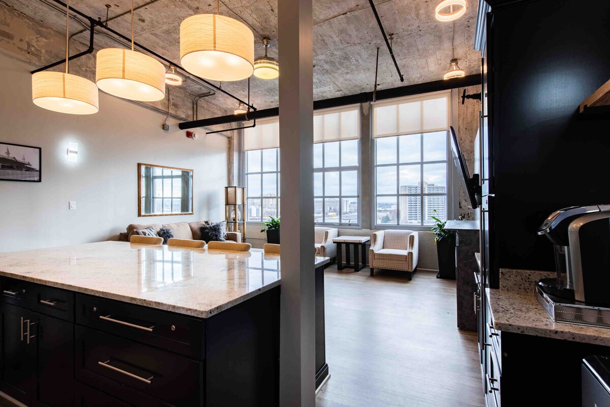 An open-plan living space features large windows allowing natural light to fill the room. A modern kitchen island with a granite countertop is visible in the foreground, while a comfortable seating area is arranged nearby, complemented by two armchairs and a cozy sofa.