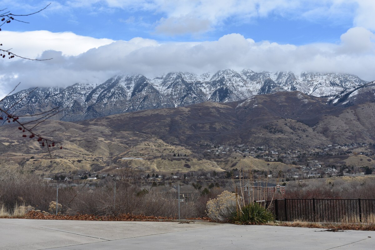 A panoramic view of snow-capped mountains is displayed under a cloudy sky, revealing varying shades of brown and green in the foreground. The landscape features a tranquil atmosphere, highlighting the natural beauty of the surrounding environment.
