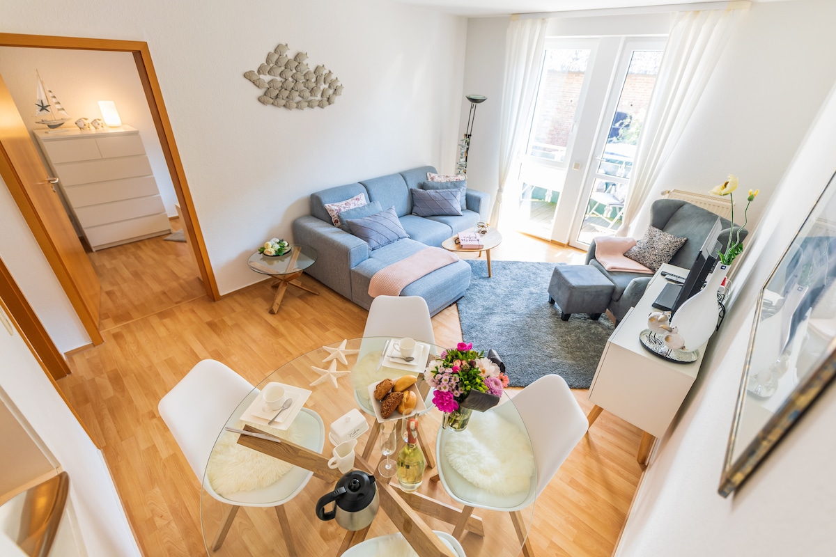 A spacious living area is featured, highlighting a comfortable gray sofa and a separate armchair positioned around a round glass dining table. Natural light streams through large glass doors leading to the balcony. Decorative elements, including a floral centerpiece, enhance the welcoming ambiance.