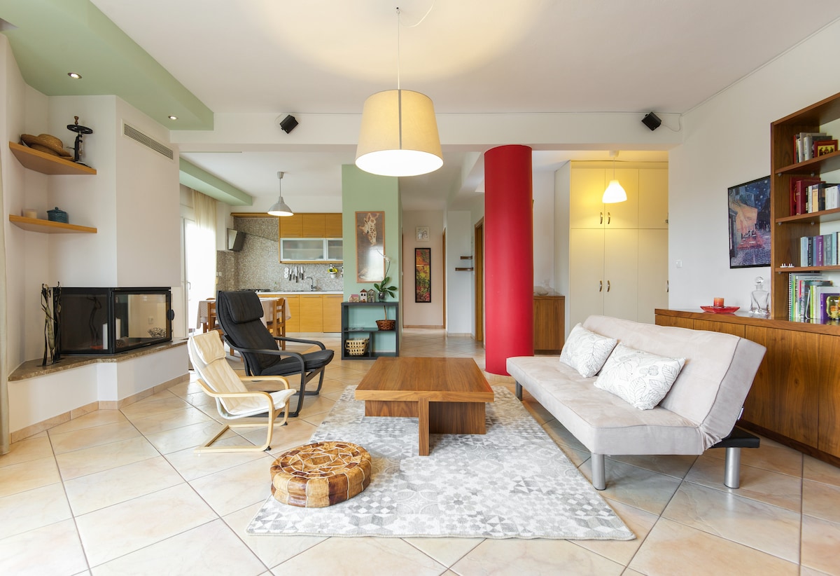 An open living space features a comfortable sofa and armchairs arranged around a central wooden coffee table. A cozy rug is underfoot, with light streaming in from large windows. A modern fireplace and unique shelving units add to the inviting ambiance.