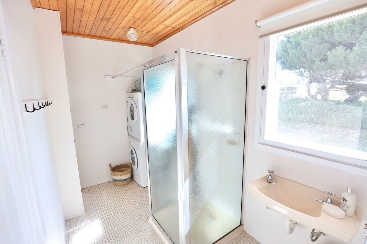 The bathroom features a glass-enclosed shower and a sink with a large mirror above. A washer and dryer unit is located to the left, while a window provides natural light and a view of the surrounding greenery. Neutral tones enhance the spacious feel.