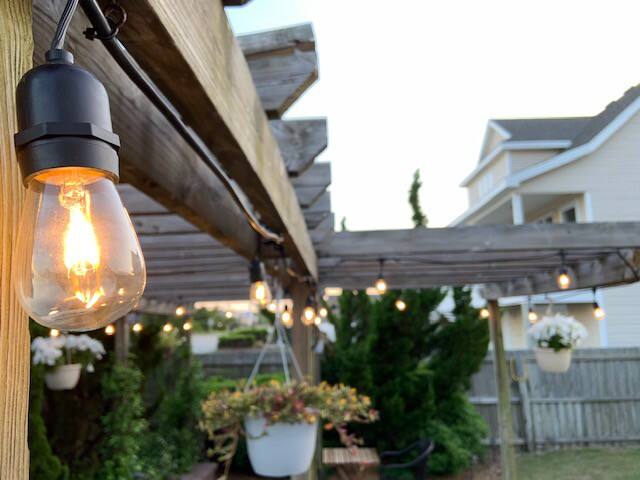 A wooden pergola is illuminated with hanging lights, creating a serene outdoor space. Potted plants hang from the beams, while additional soft lighting accentuates the lush greenery in the background. The setting presents a welcoming atmosphere for relaxation.