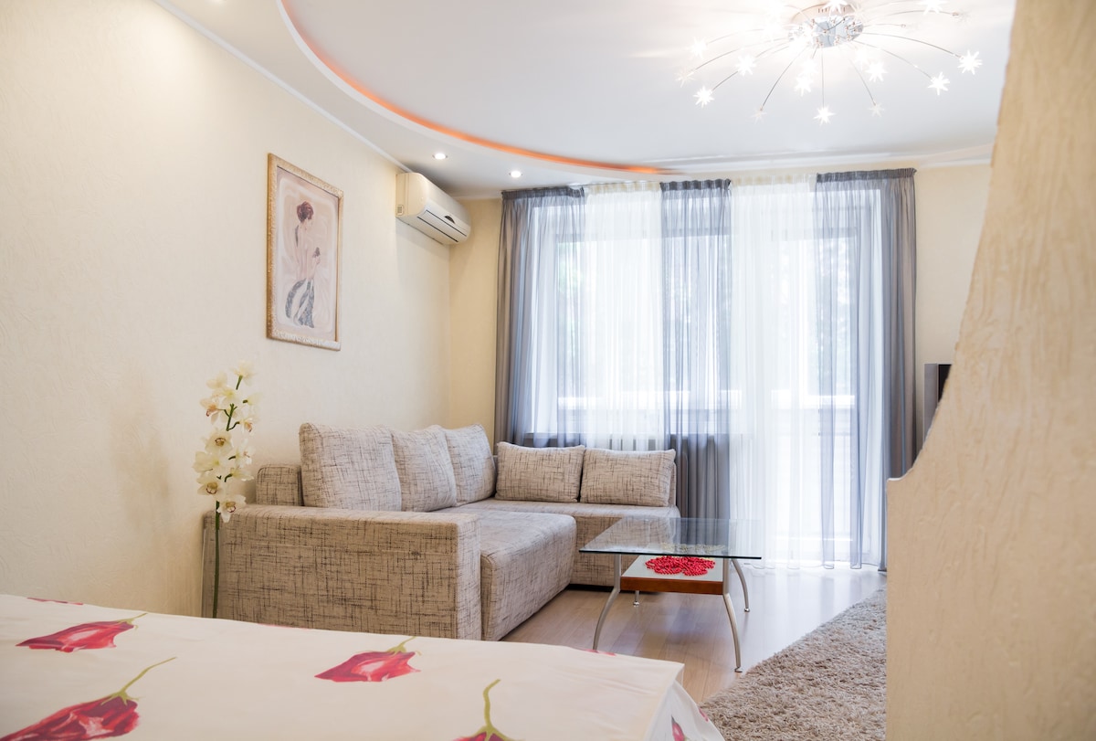 A light-filled living area features a modern gray sectional sofa accompanied by a glass coffee table. Sheer curtains frame the window, allowing soft natural light to filter in. A decorative wall art piece and a floral arrangement add subtle touches to the space.