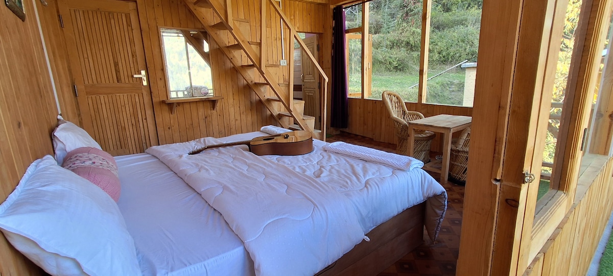 A wooden interior features a spacious bed with white bedding, accompanied by a guitar resting on top. Large windows allow natural light to fill the room, showcasing a rustic staircase leading to a lofted area. A small table and chairs are visible near the window.