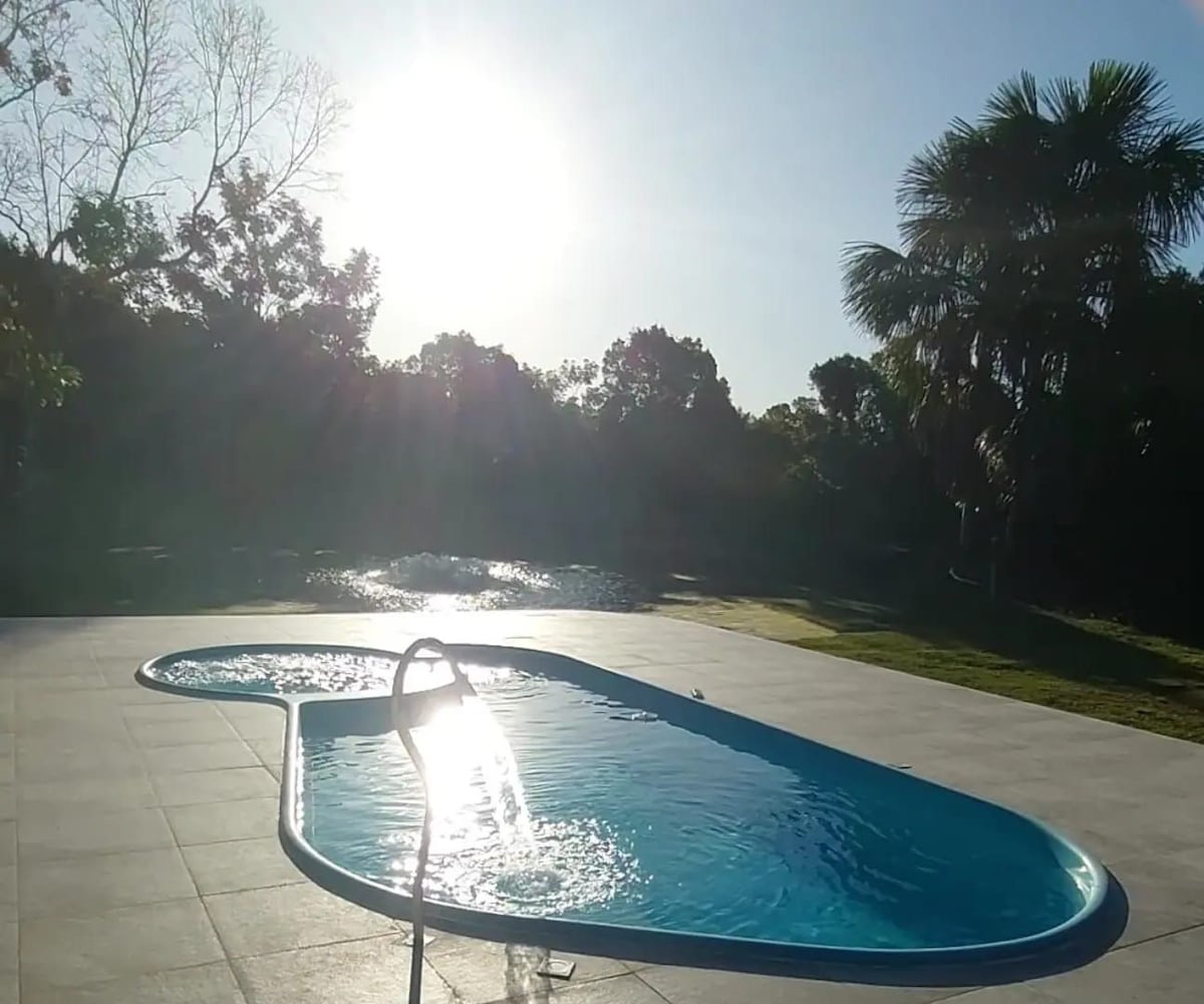 A swimming pool is seen with a curved shape, reflecting sunlight. Surrounding vegetation includes trees and palm plants, adding to the natural ambiance. The pool area features tiled flooring and is situated near a tranquil body of water.