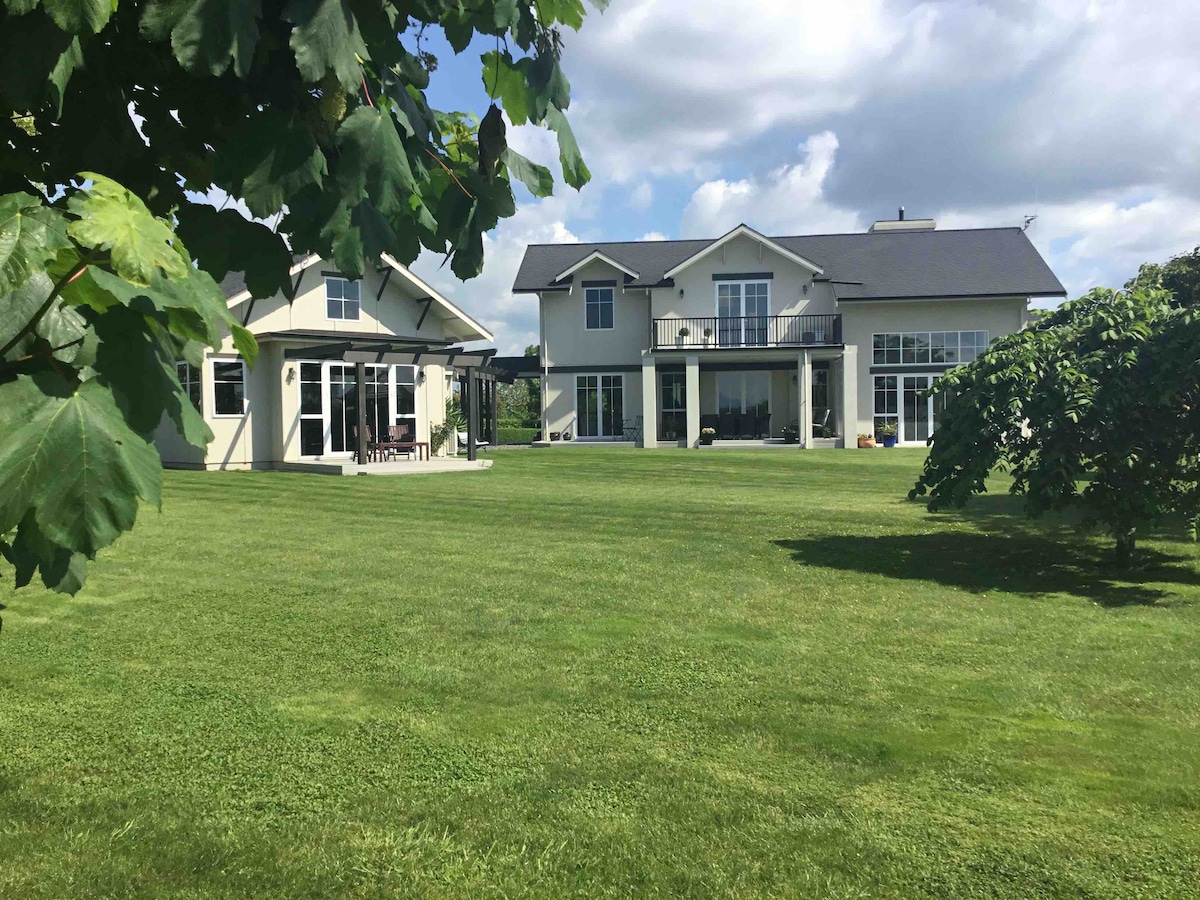 Two spacious cottages are set on a well-maintained lawn, surrounded by greenery. Large windows allow for ample natural light, with outdoor seating visible on the deck. The expansive sky features soft, fluffy clouds, contributing to a serene countryside atmosphere.