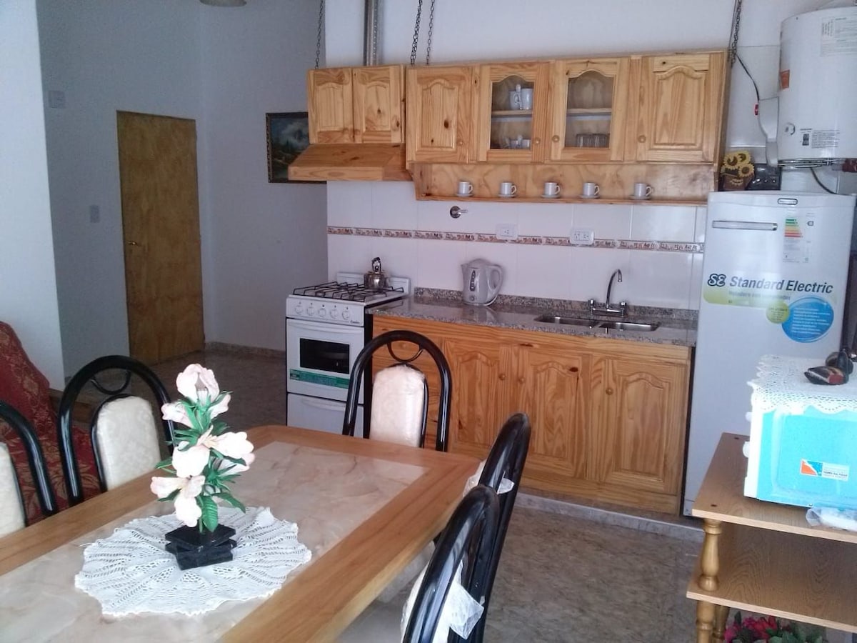 The kitchen area features wooden cabinetry and a counter with a sink and a stove. A dining table with black chairs is positioned nearby, adorned with a decorative white tablecloth and a vase of flowers. Appliances include a refrigerator and a kettle.