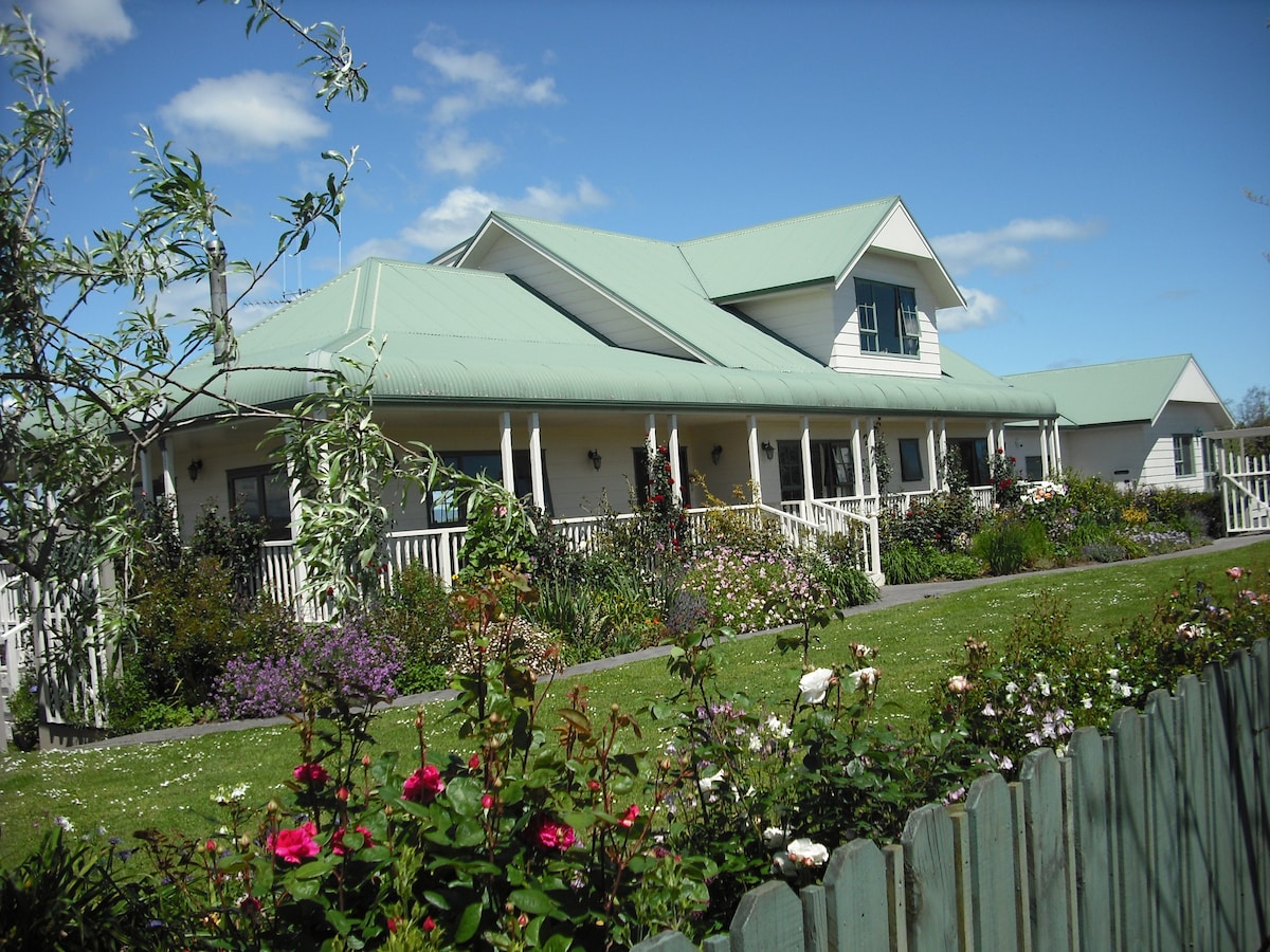 A spacious two-story house with a green tin roof is set amidst colorful flower gardens. Surrounding greenery and a picket fence enhance the home's charming exterior, under a blue sky adorned with fluffy clouds.