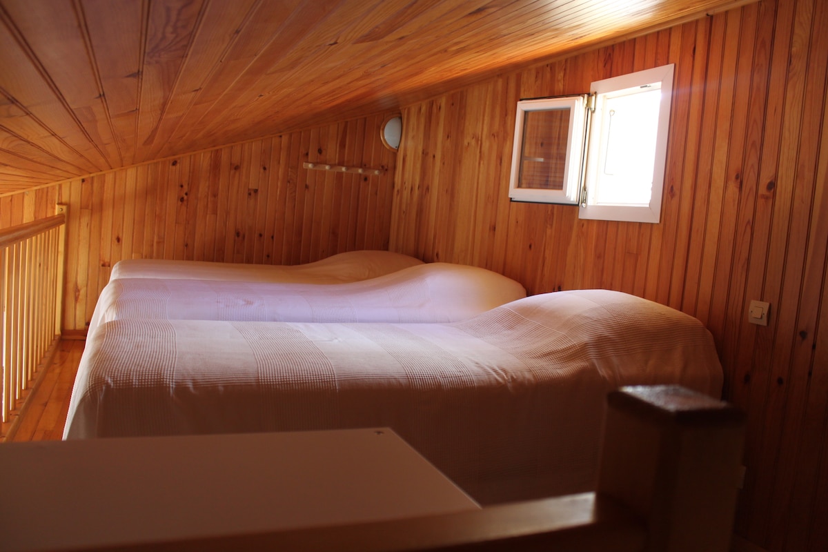 The mezzanine area features two spacious beds, each covered with light-colored linens. The natural wood paneling creates a warm environment, complemented by a small window allowing natural light to fill the space.