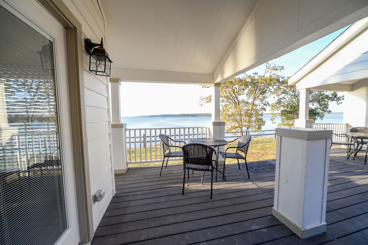 A semi-private deck is shown, featuring a round table with four black chairs. The deck overlooks tranquil water views, framed by trees. Soft natural light enhances the inviting atmosphere of the outdoor space, perfect for relaxing or dining.