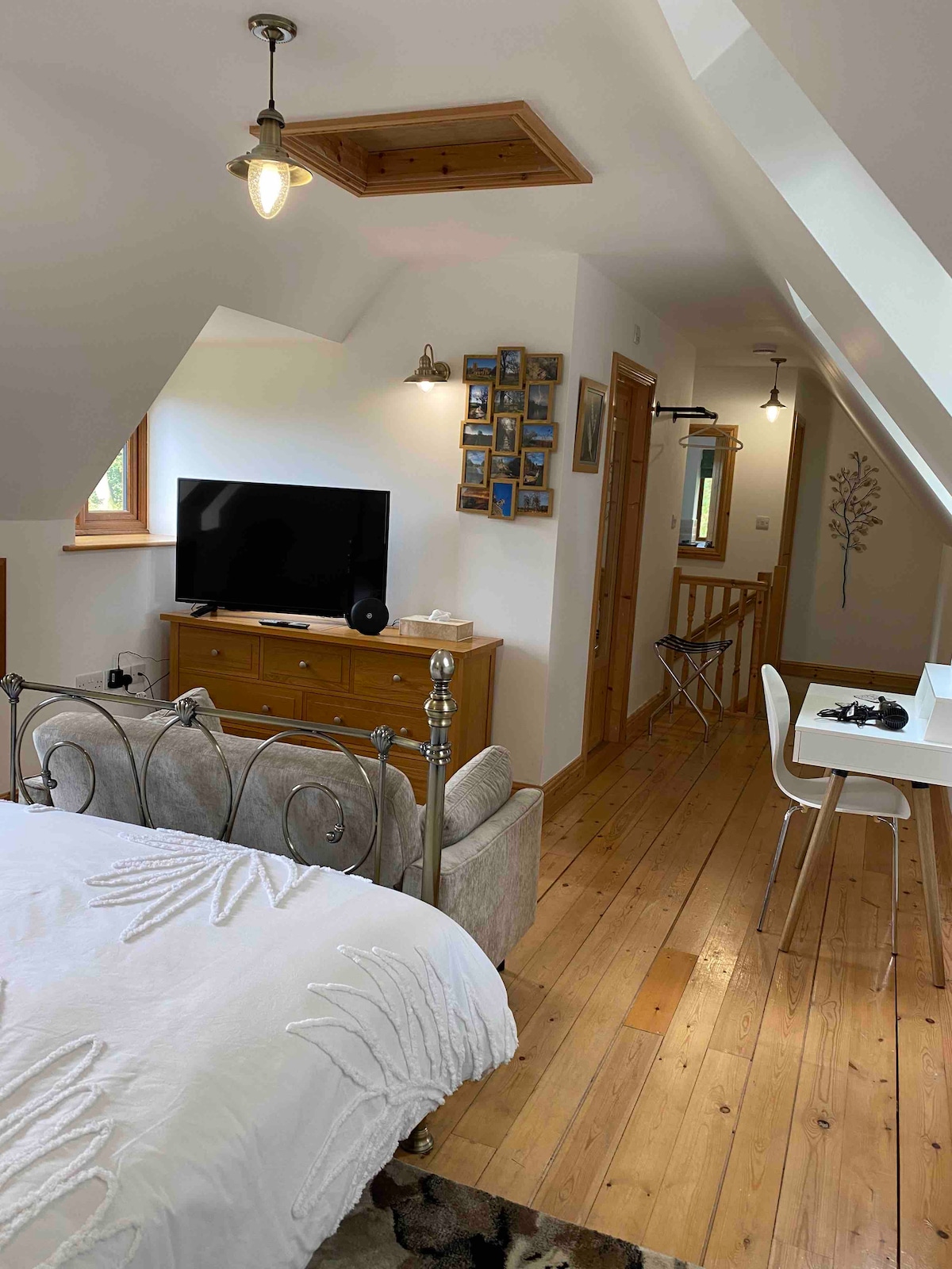 A cozy space is presented with a comfortable bed featuring a light-colored blanket. A wooden dresser and a television are positioned in front. Light streams in from windows, enhancing the polished wooden floor and highlighting the open layout towards a small dining area.