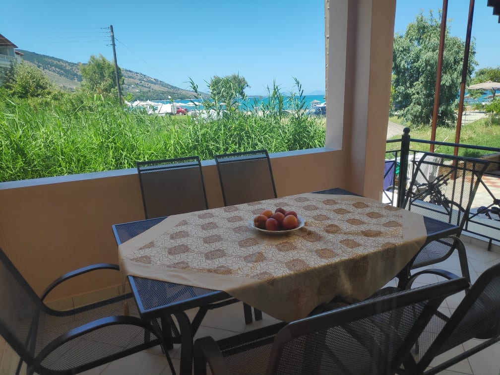 A dining table, set with a decorative cloth and a bowl of fruit, is situated on a covered patio. Greenery and a view of the Ionian Sea are visible in the background, enhancing the inviting outdoor space.