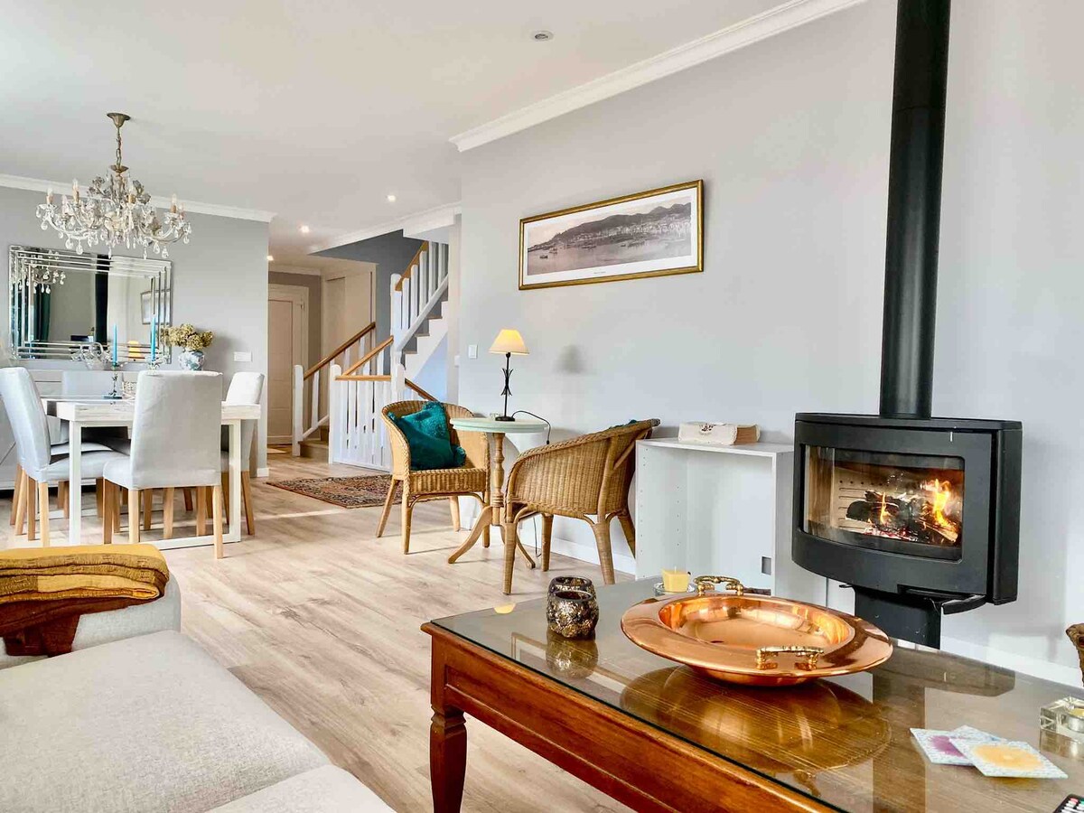 A warm and inviting living space features a comfortable seating area with a sofa and two wicker chairs. A coffee table with a decorative tray is positioned in front of a modern fireplace, while a dining area with a chandelier is visible in the background, creating a cozy atmosphere.