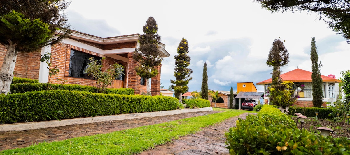 A well-maintained pathway leads to a brick-fronted building, flanked by neatly trimmed hedges and trees. The landscape features a spacious green lawn and additional buildings with red roofs visible in the background, under a partly cloudy sky.