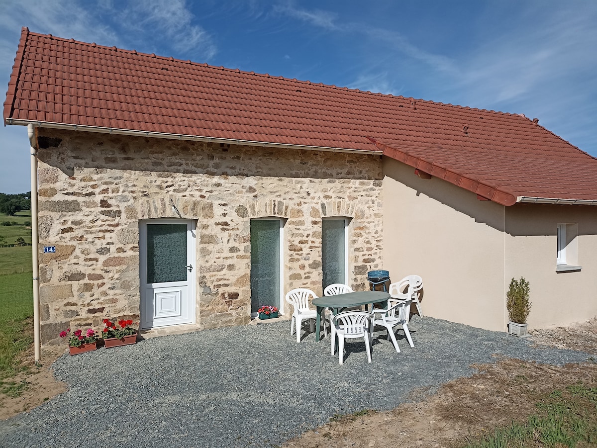 The exterior of the house is shown, featuring a rustic stone facade and a red-tiled roof. A small patio area is visible, furnished with a table and six white plastic chairs. The surrounding landscape includes greenery, and a clear blue sky is above.