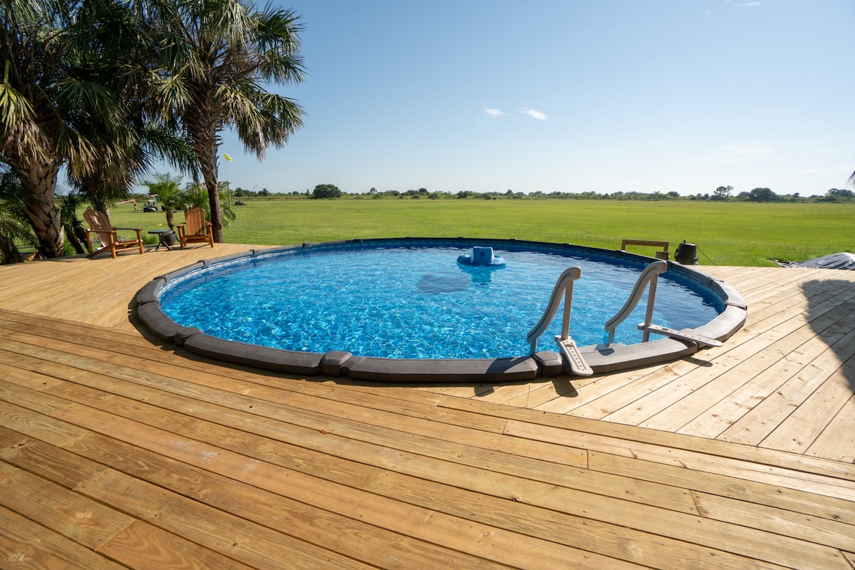 A circular swimming pool is surrounded by wooden decking and palm trees, offering a serene view of the open landscape. A ladder provides access to the water, and a built-in seating area is visible in the center of the pool.