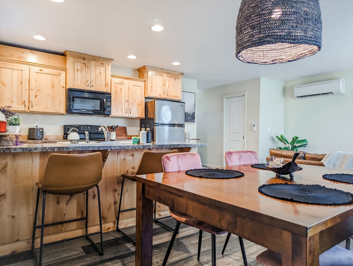 A modern kitchen is featured with wood cabinetry and stainless steel appliances. An island with seating for three is complemented by a dining table that accommodates four. A woven pendant light adds texture to the space, enhancing the inviting kitchen atmosphere.