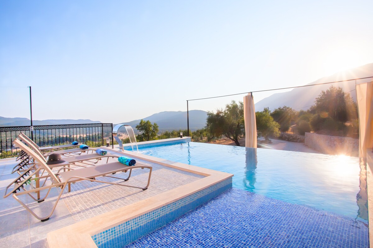 The image features a private infinity pool with lounging chairs, overlooking a scenic landscape. The pool reflects the tranquil blue sky and surrounding hills, while the sun casts a warm glow across the area, providing a serene backdrop for relaxation.