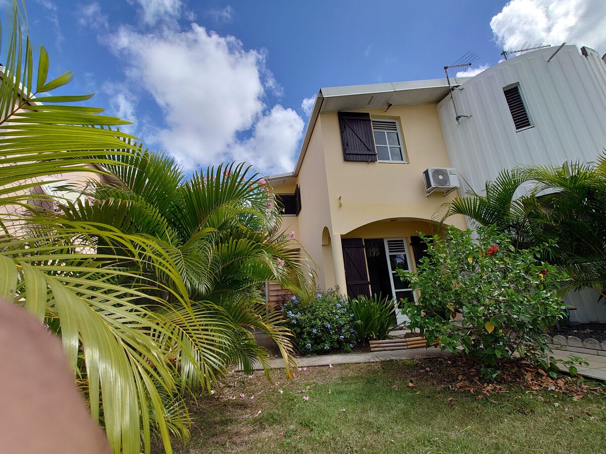 The exterior of the house is framed by lush green plants and palm trees. The two-story structure features light yellow walls and dark wooden shutters on the windows. A cooling unit is visible on the side, complementing the tropical setting under a clear blue sky.