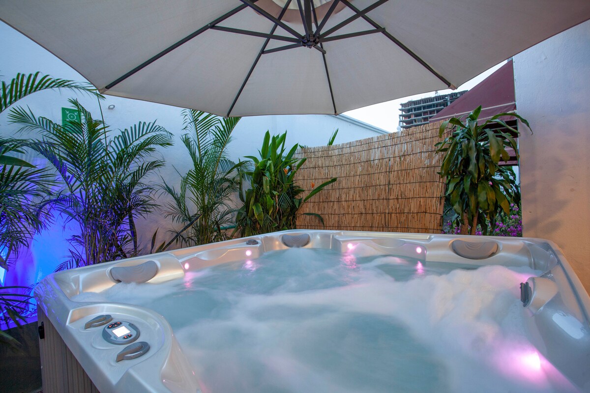 A private hot tub is surrounded by lush greenery and bamboo fencing, offering a secluded relaxation space. Soft lighting enhances the atmosphere, while a large umbrella provides shade overhead.