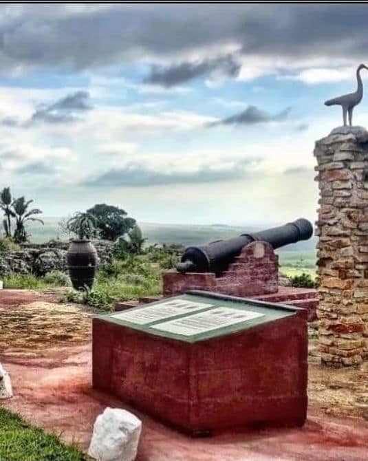 An outdoor historical site features an ancient cannon positioned on a stone platform, surrounded by lush greenery and open skies. An informative sign rests on a flat, rectangular stone, providing details about the site. In the background, a distant landscape is visible.