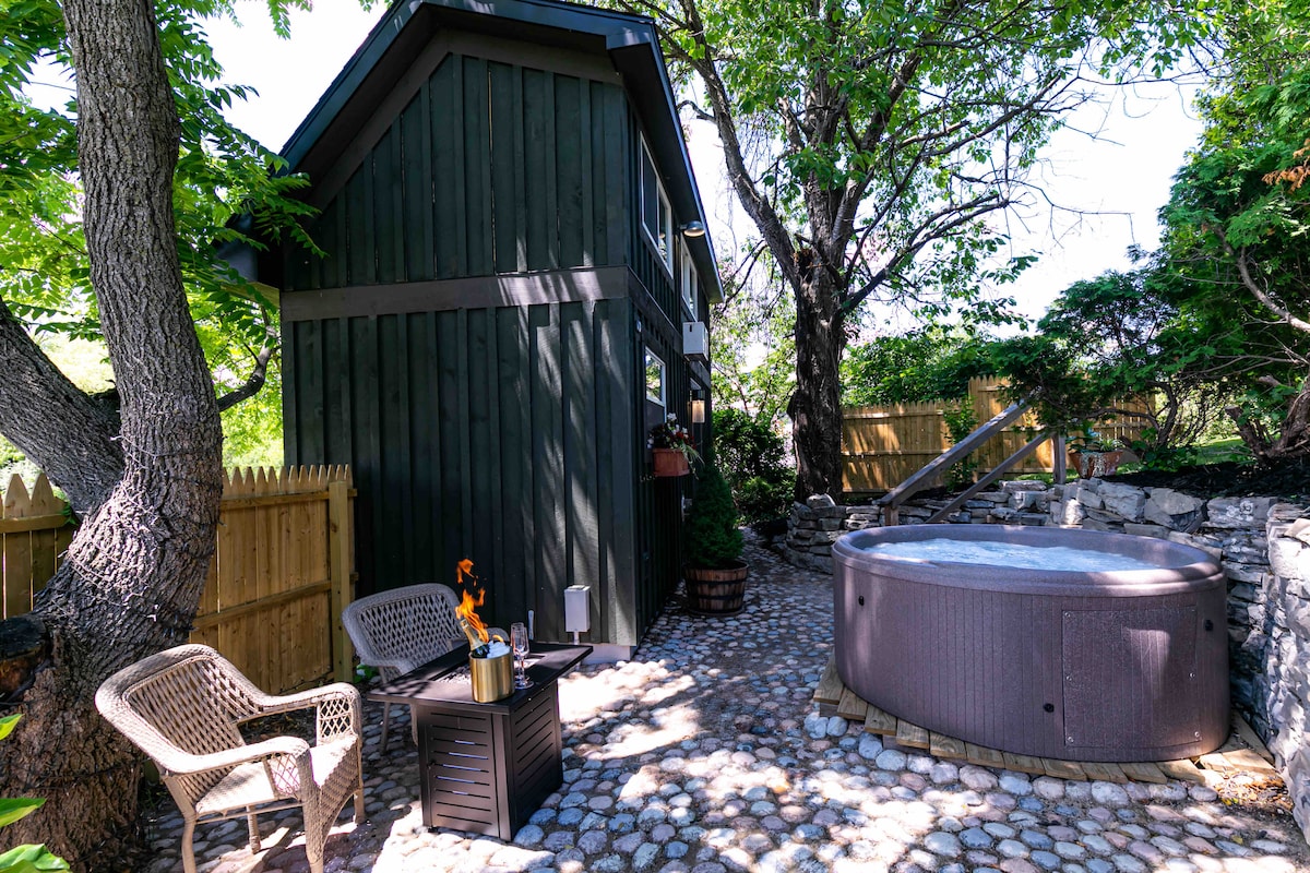 An outdoor area surrounding a tiny home features a private hot tub surrounded by stone pavers. Two cozy chairs are positioned near a small table, creating an inviting space for relaxation. Lush greenery and trees provide shade and privacy in the landscaped setting.