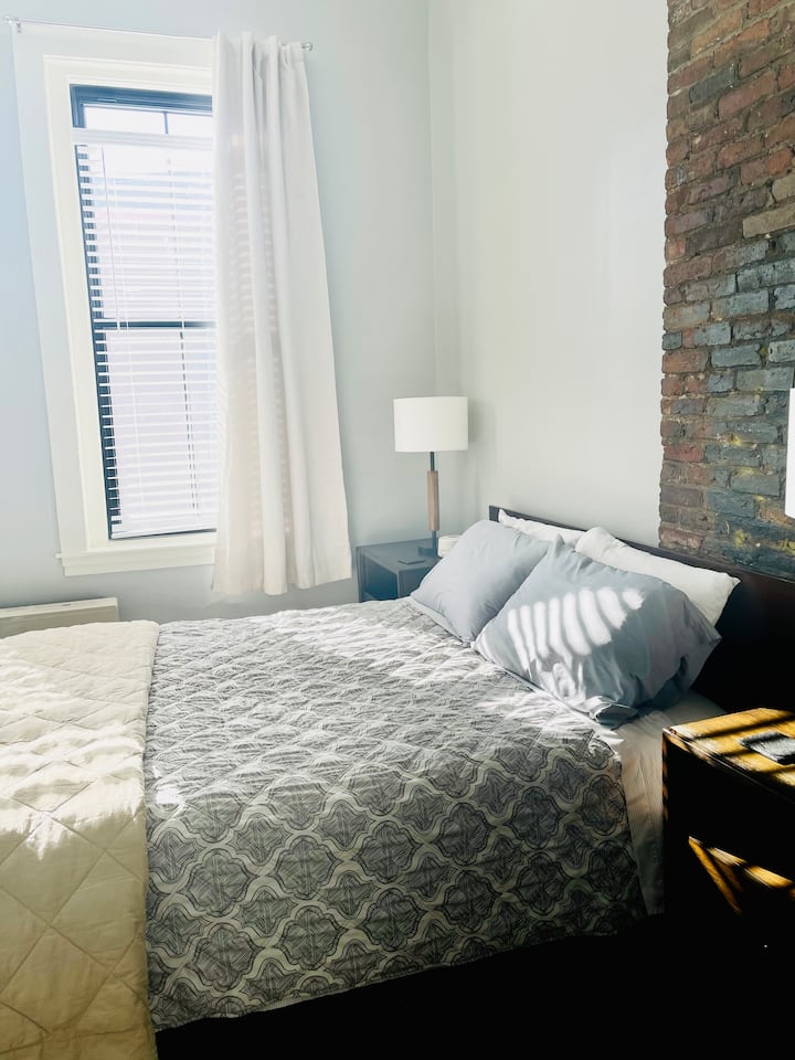 Chumba cha kulala kilicho na kitanda cha malkia, bandari ya kuchaji, mapazia ya kuongeza giza kwenye chumba, na mashine ya kupiga kelele nyeupe.