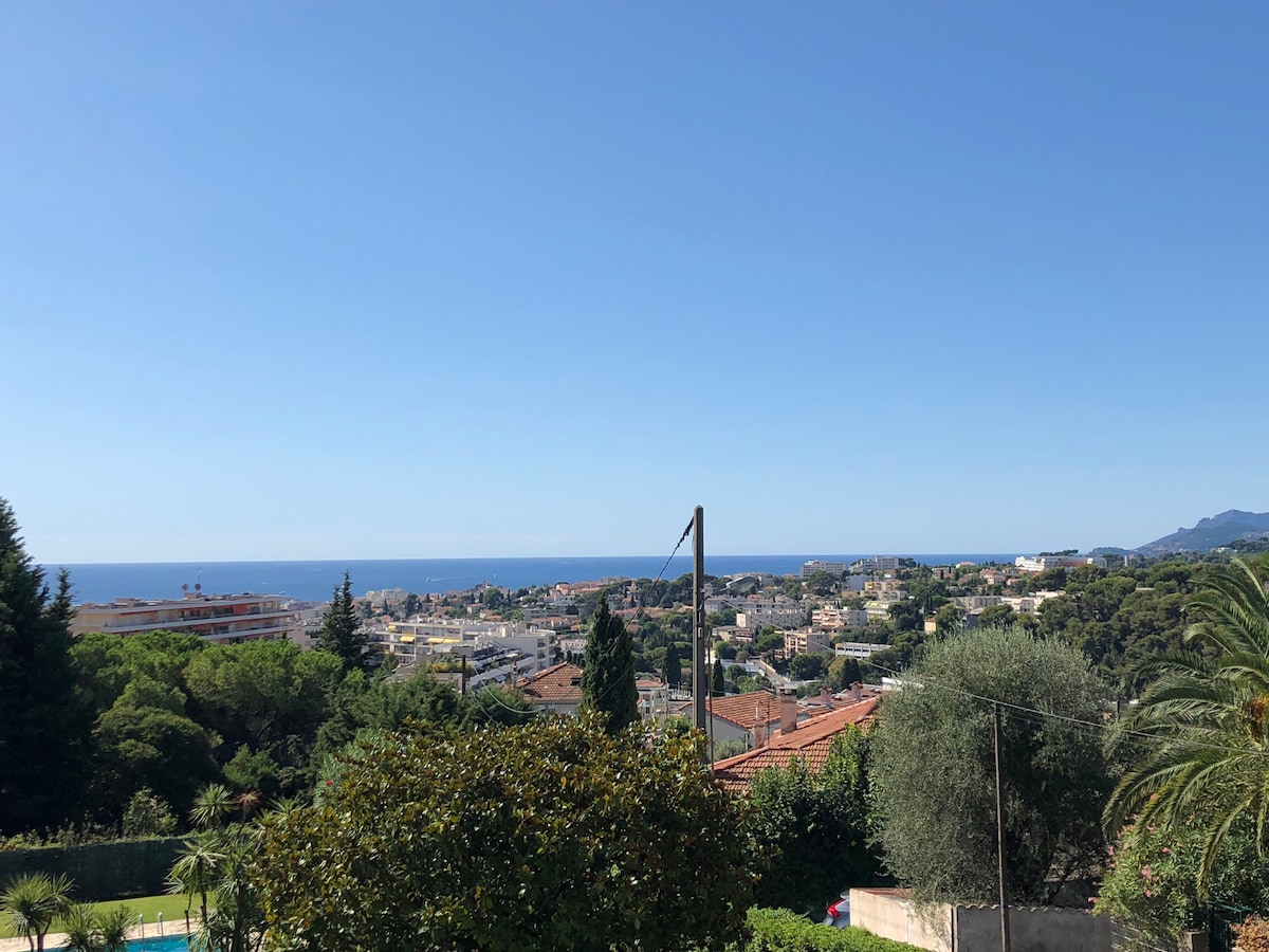 A panoramic view showcases the blue sea in the distance alongside the greenery of surrounding trees and well-maintained gardens. Buildings and homes can be seen below, set against a clear blue sky that enhances the tranquil atmosphere of the area.
