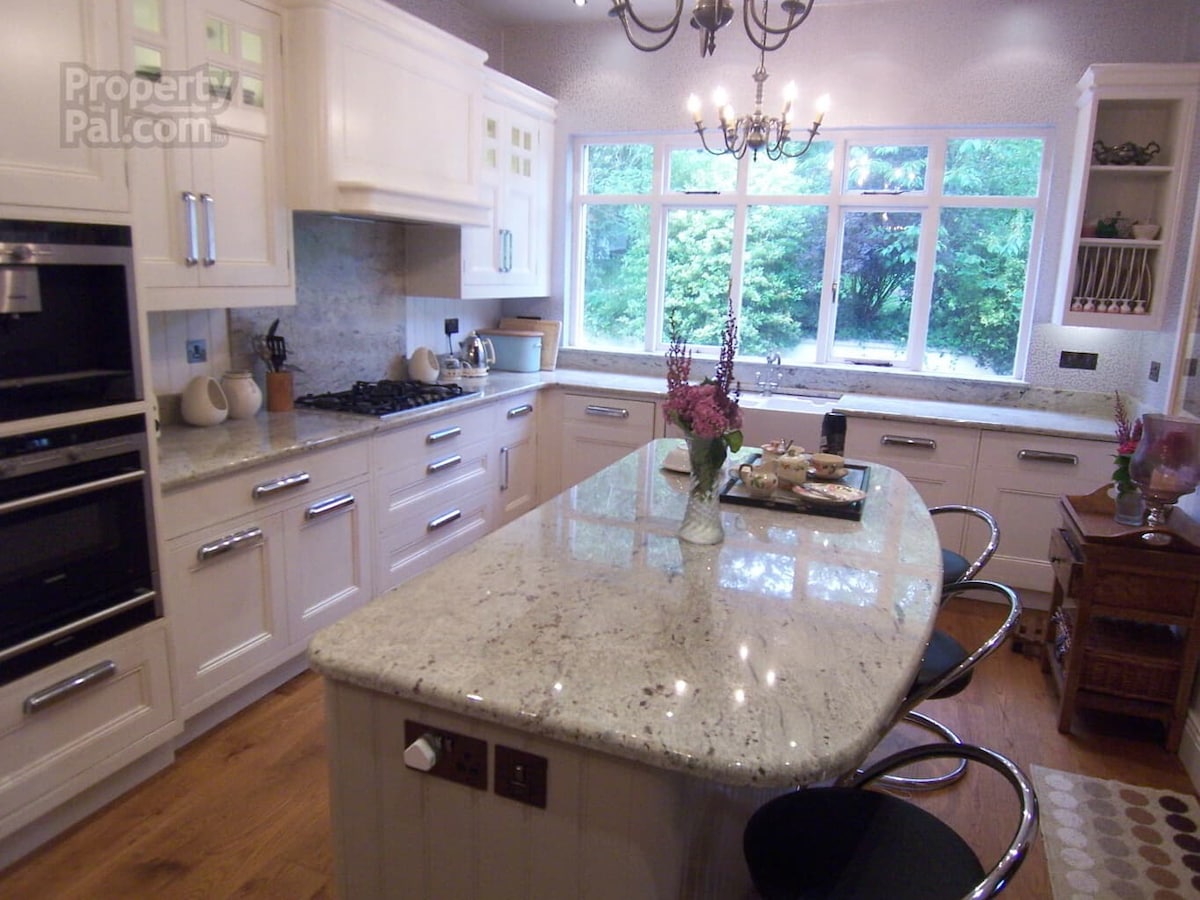 A modern kitchen features a central granite island with seating and an array of kitchen appliances. Natural light streams in through large windows, highlighting the cabinetry and countertops. A subtle decorative arrangement adds a welcoming touch to the space.