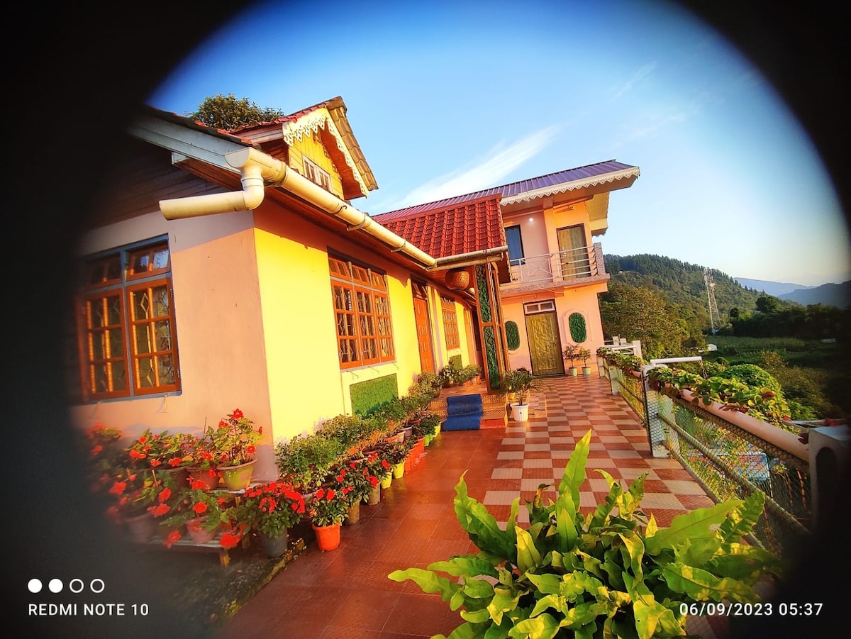 The exterior of a sunlit house features a tiled patio adorned with vibrant flowers in planters. Large windows allow for natural light, while a balcony extends to the side. The surrounding landscape includes rolling hills, enhancing the natural beauty of the space.