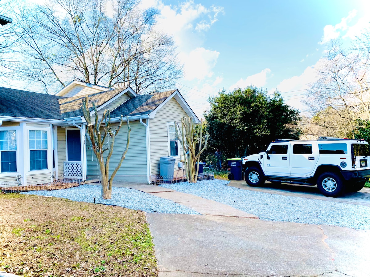 The exterior view features a charming house situated among trees, with a gravel driveway leading to a parked vehicle. A landscaped front yard is visible, complemented by a fence, enhancing the overall appeal of the property.