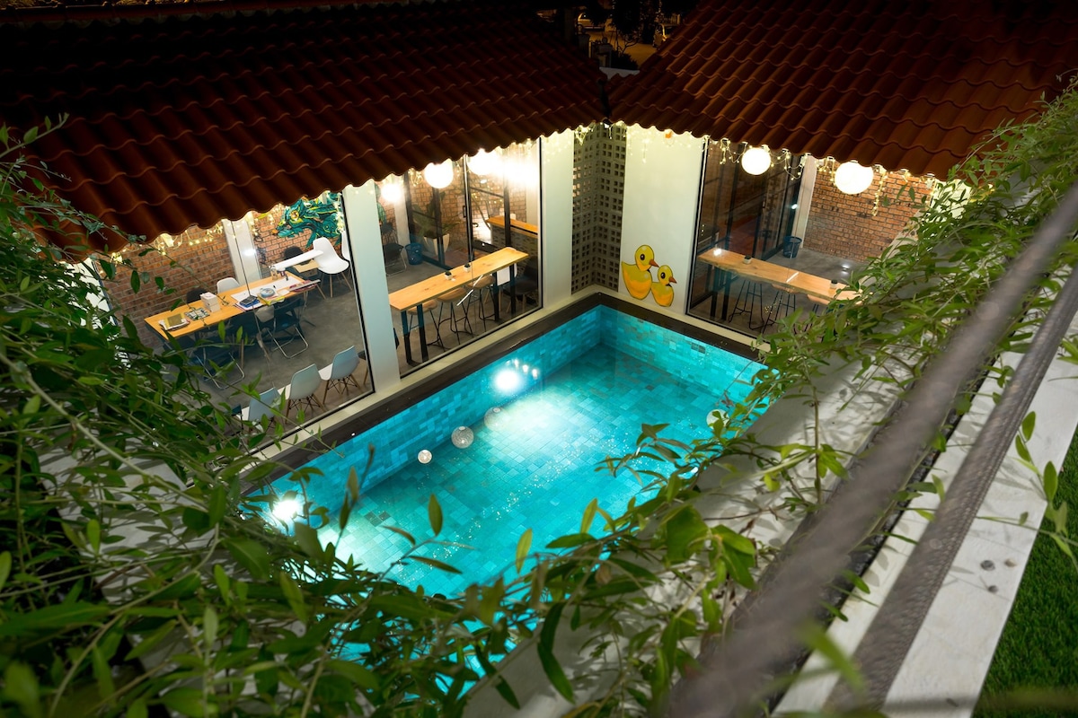 An outdoor swimming pool is surrounded by lush greenery and two structures with terracotta roofs. Tables with seating are visible near the pool area, illuminated by soft, warm lighting that enhances the evening ambiance.