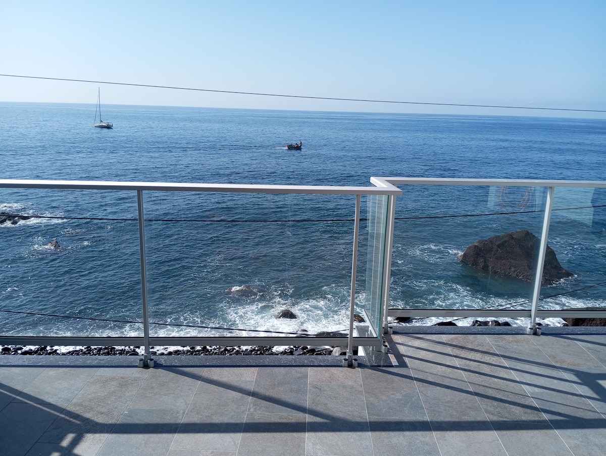 An ocean view is presented from the spacious terrace. A glass railing provides safety while framing the sparkling water below. Gentle waves meet rocky formations, with distant boats visible on the horizon under a clear blue sky.