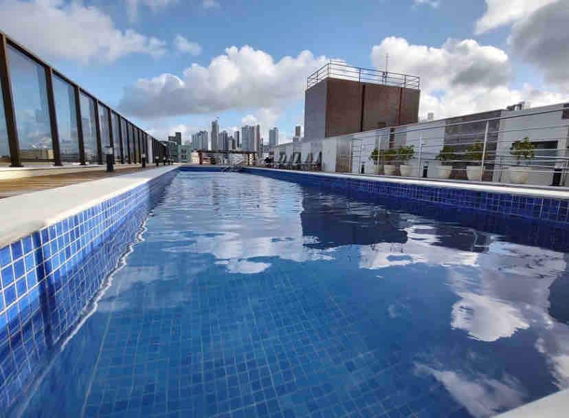 The rooftop pool features a clear blue surface reflecting the sky and surrounding skyline. Lounge chairs are positioned along the deck, providing comfortable seating. A few potted plants add natural elements to the modern space, enhancing the pool area.