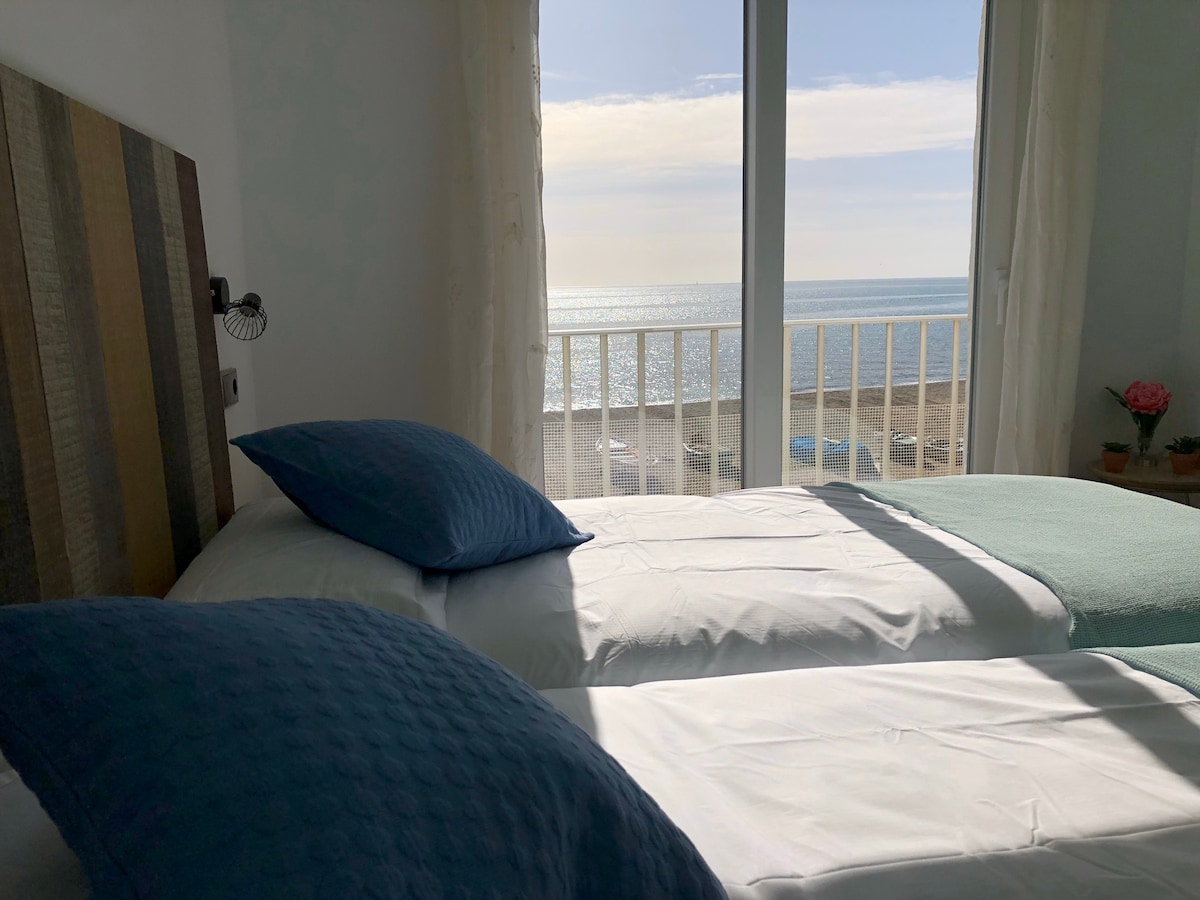 A bright sleeping area features two spacious beds adorned with blue pillows, positioned beside large windows that offer views of the sea. Soft natural light fills the room, and a small potted plant sits on the windowsill, adding a touch of color.