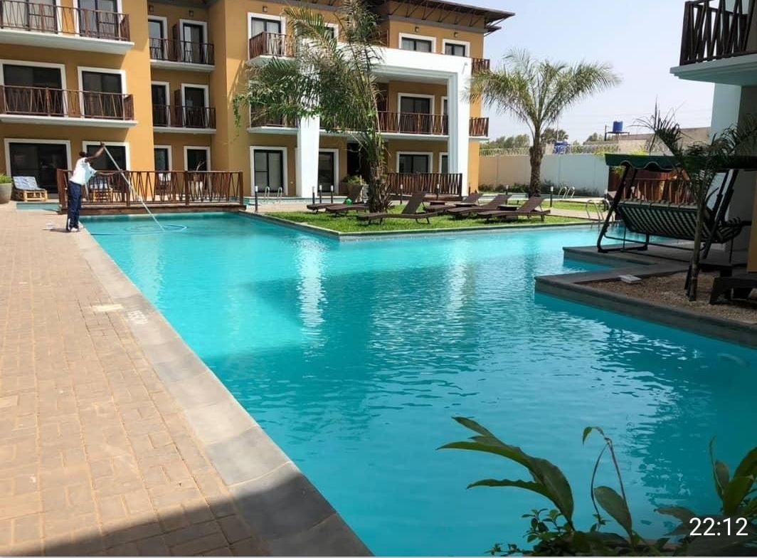 A refreshing pool area is flanked by lounge chairs and palm trees, set against a backdrop of a multi-story building. The water is clear and inviting, with a maintenance staff member ensuring cleanliness. The surrounding landscaping features green grass and decorative stone accents.