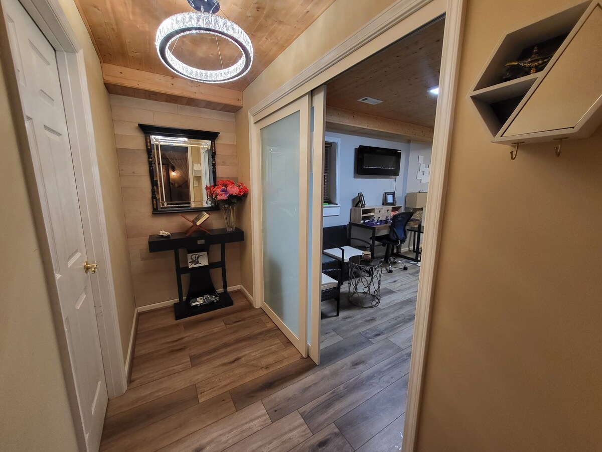 A well-lit entryway features wooden flooring and neutral walls. A stylish mirror is mounted above a console table adorned with flowers. Glass doors lead to an office space, which contains a desk and multiple chairs, enhancing the functional layout.