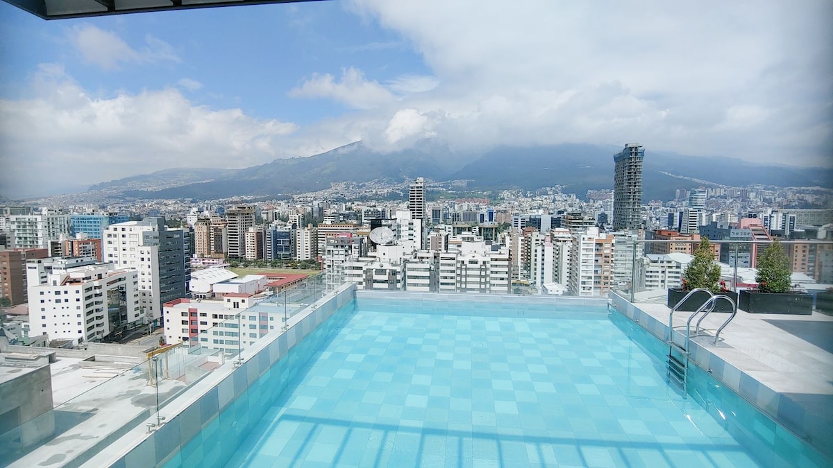 A rooftop infinity pool offers a clear view of the city and mountains beyond. The serene water reflects the sky and surrounding buildings, creating a tranquil atmosphere. Rooftop amenities are visible, encouraging relaxation and enjoyment of the cityscape.
